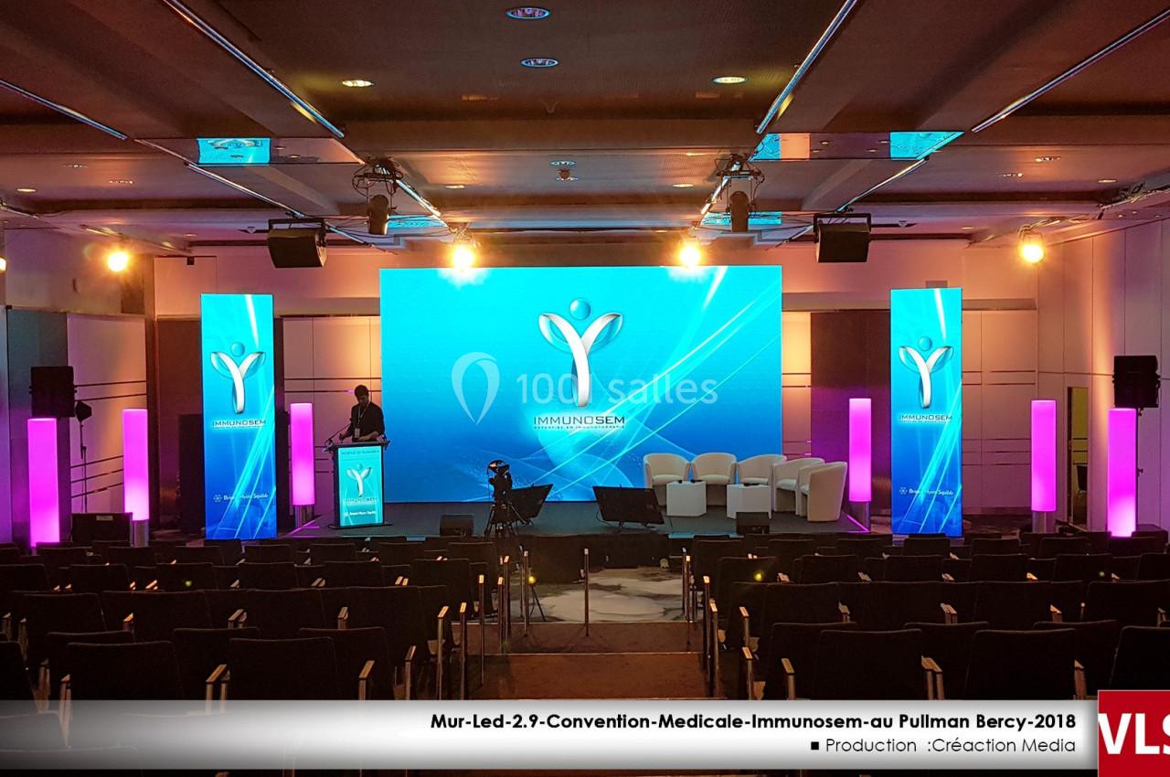 Salle de conférence avec scène équipée d'écrans lumineux, fauteuils blancs et projecteurs, prête pour un événement.