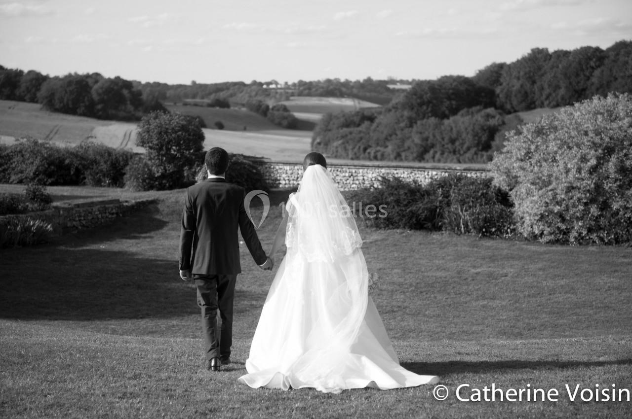 Un couple de mariés marche main dans la main dans un jardin, avec une vue dégagée sur la campagne.