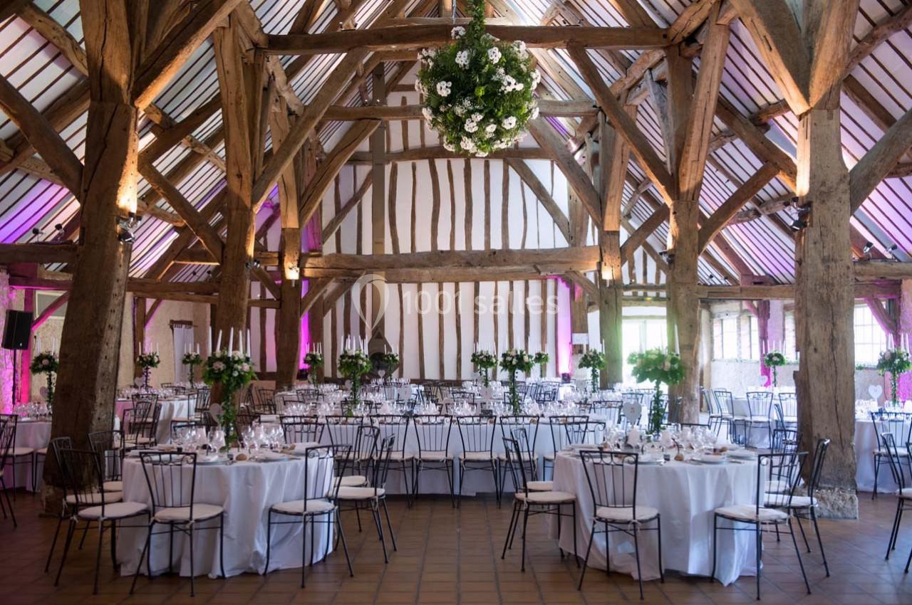 Salle de réception rustique avec charpente en bois, tables rondes dressées et décoration florale suspendue.