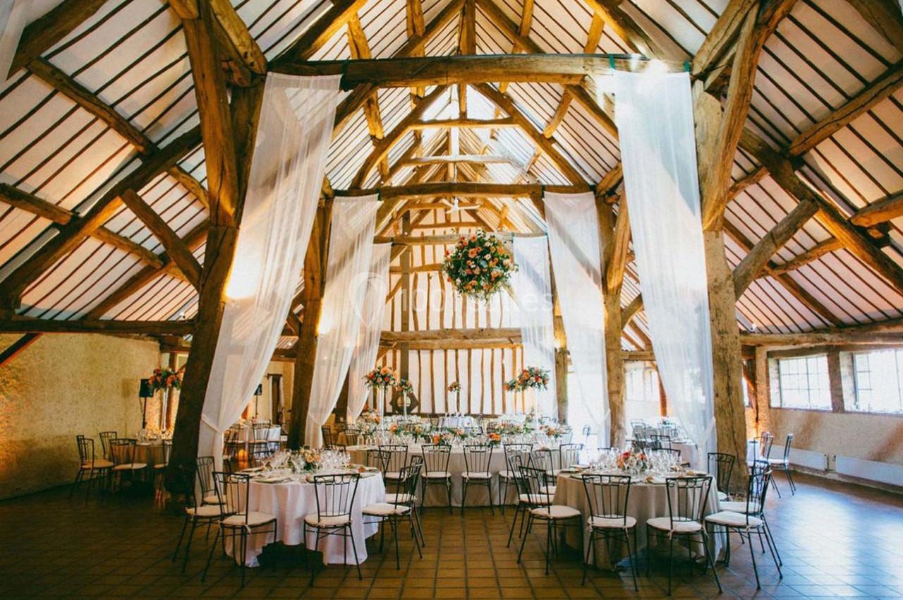 Salle de réception avec poutres apparentes, tables rondes dressées et décoration florale suspendue.