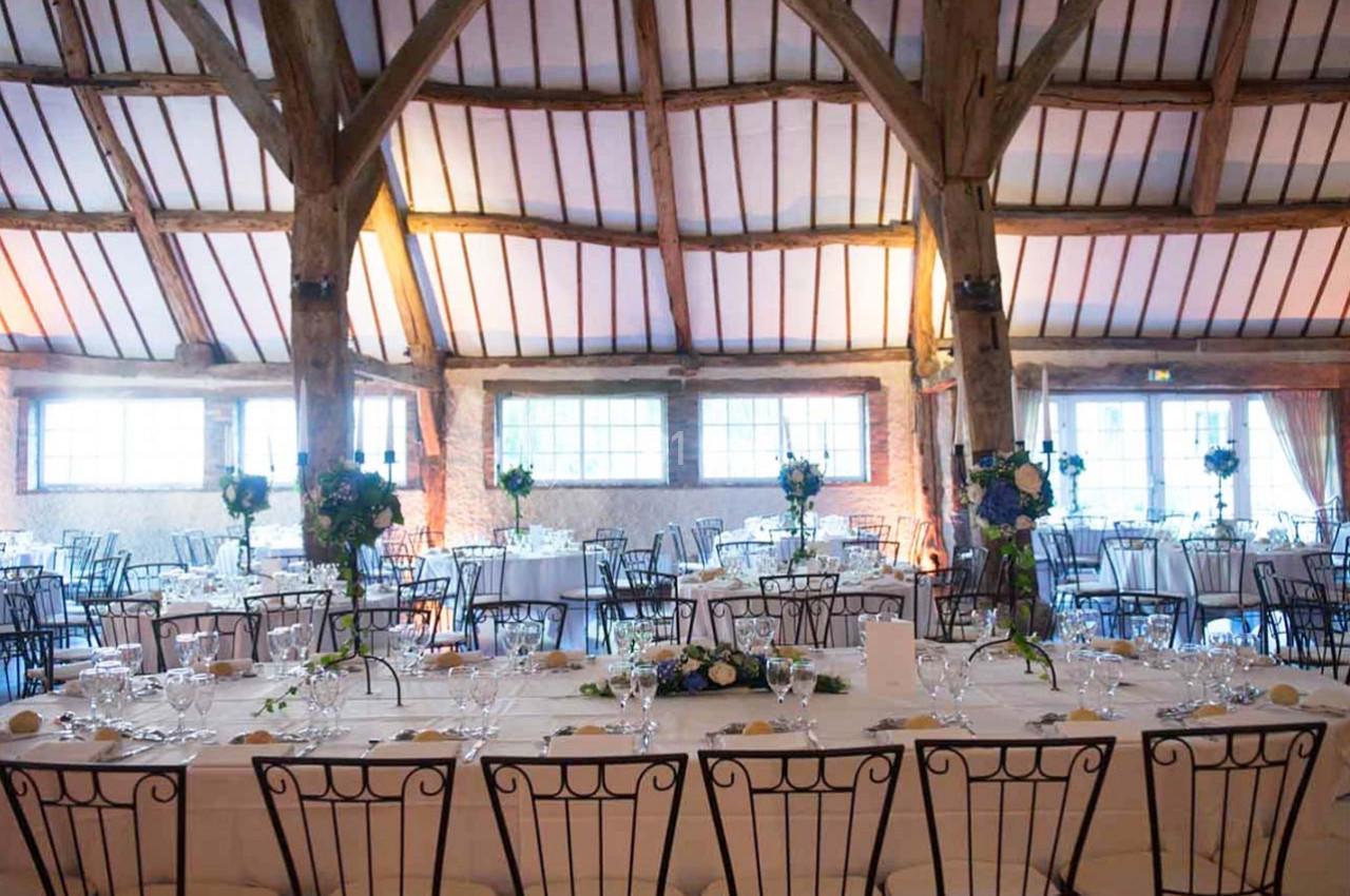 Salle de réception avec tables dressées, chaises en métal, poutres apparentes et grandes fenêtres laissant entrer la lumière.
