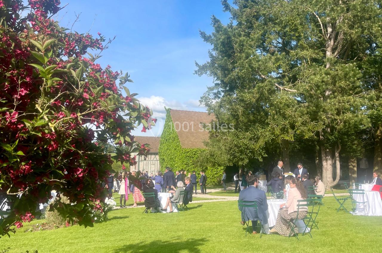 Personnes rassemblées dans un jardin verdoyant avec des tables, des arbres et un bâtiment en arrière-plan par temps…