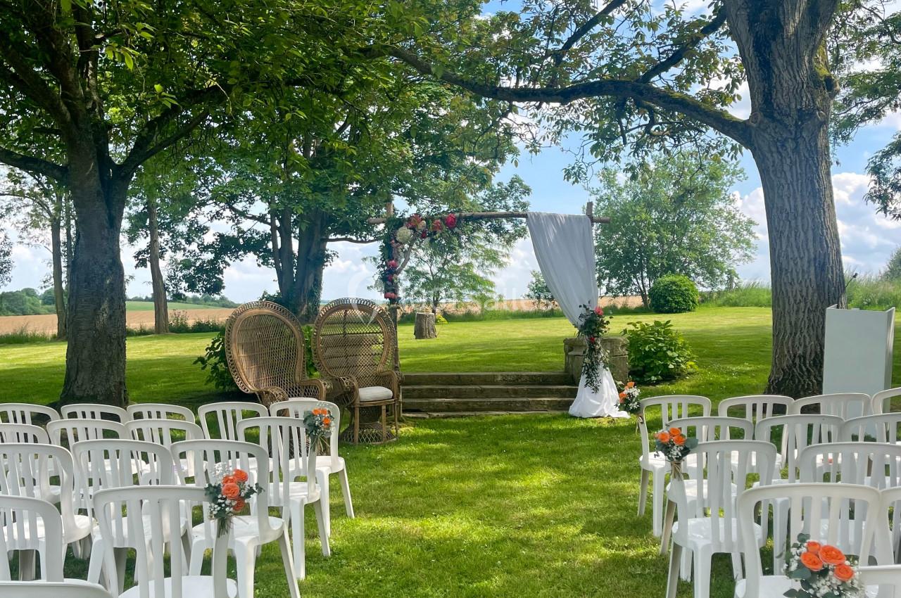 Chaises blanches alignées en extérieur face à une arche décorée, sous des arbres dans un cadre verdoyant.