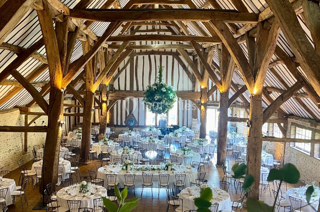 Grande salle de réception avec charpente en bois apparente, tables rondes dressées et décoration florale suspendue.