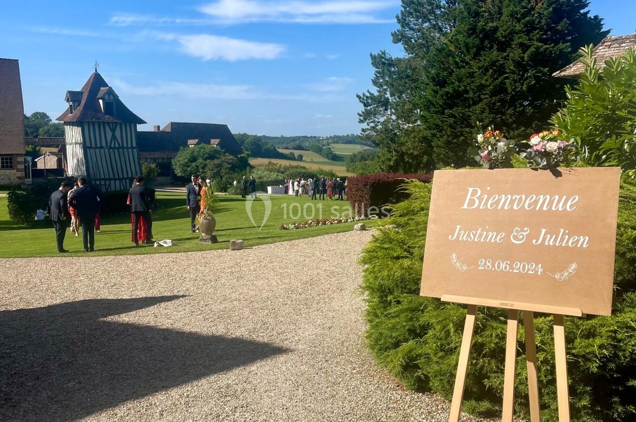 Panneau de bienvenue pour un mariage, placé sur un chevalet dans un jardin avec des invités en arrière-plan.