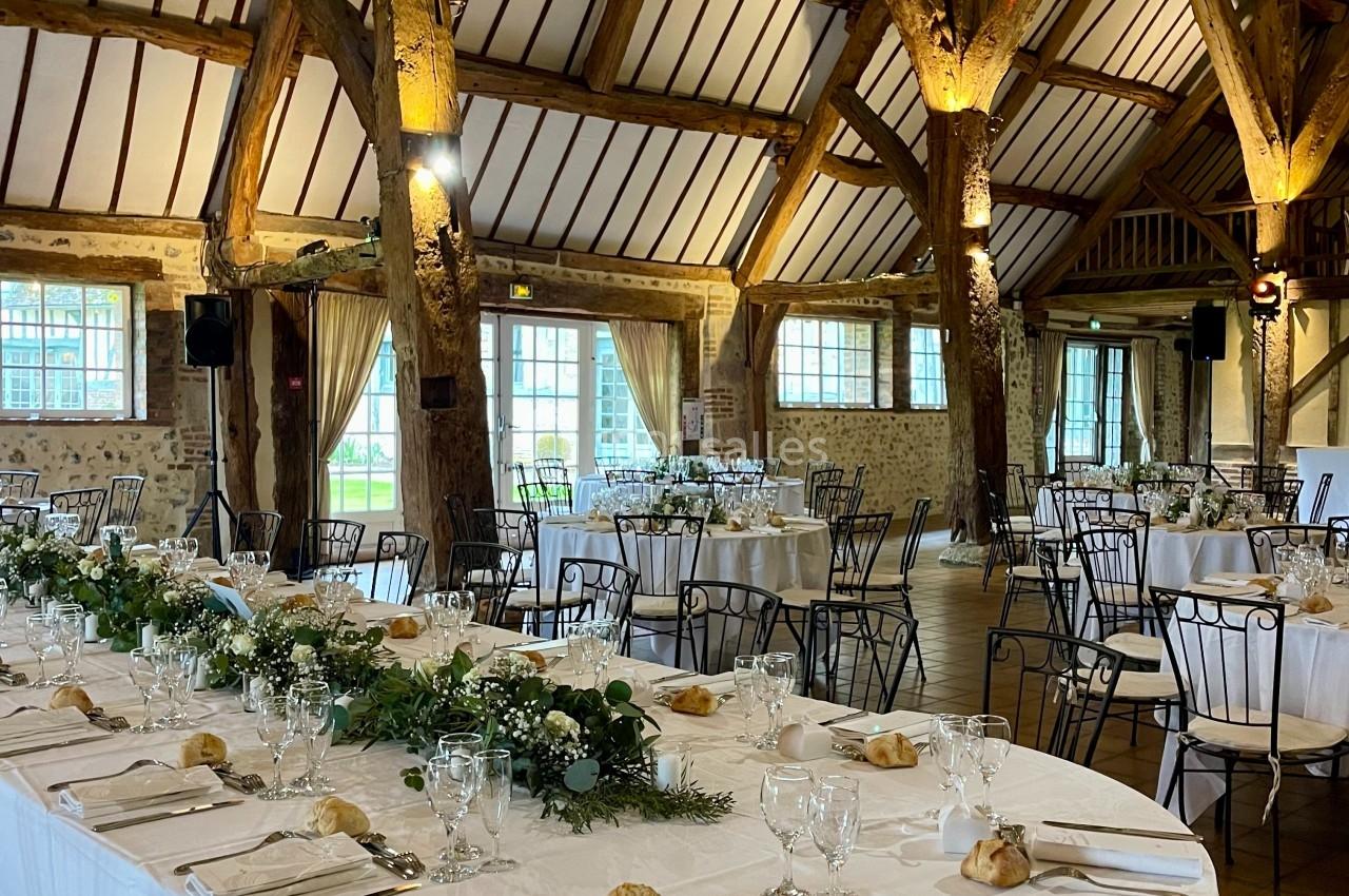 Salle de réception avec charpente en bois, tables dressées avec nappes blanches, vaisselle élégante et décoration florale.