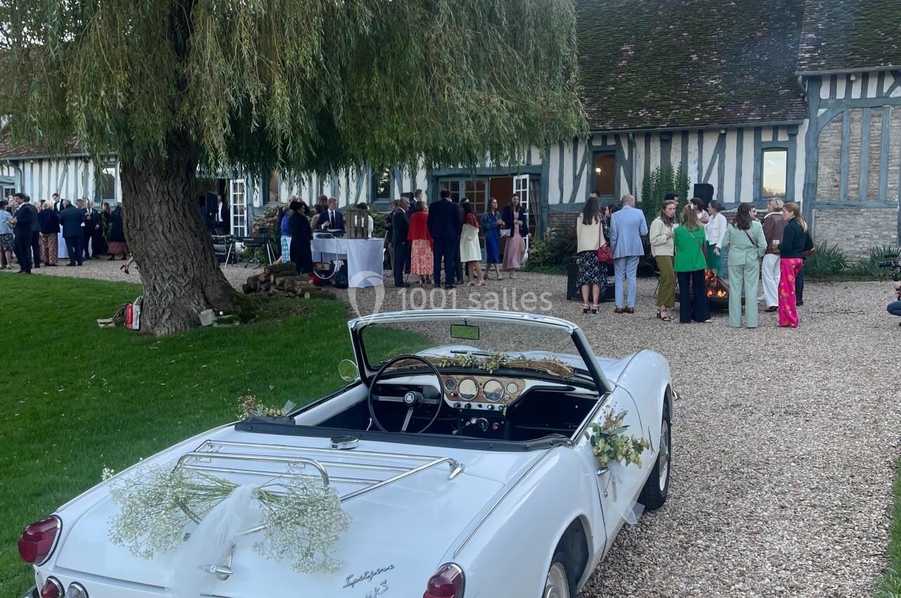 Voiture décapotable blanche décorée pour un mariage, stationnée devant une maison à colombages avec des invités à l'arrière…