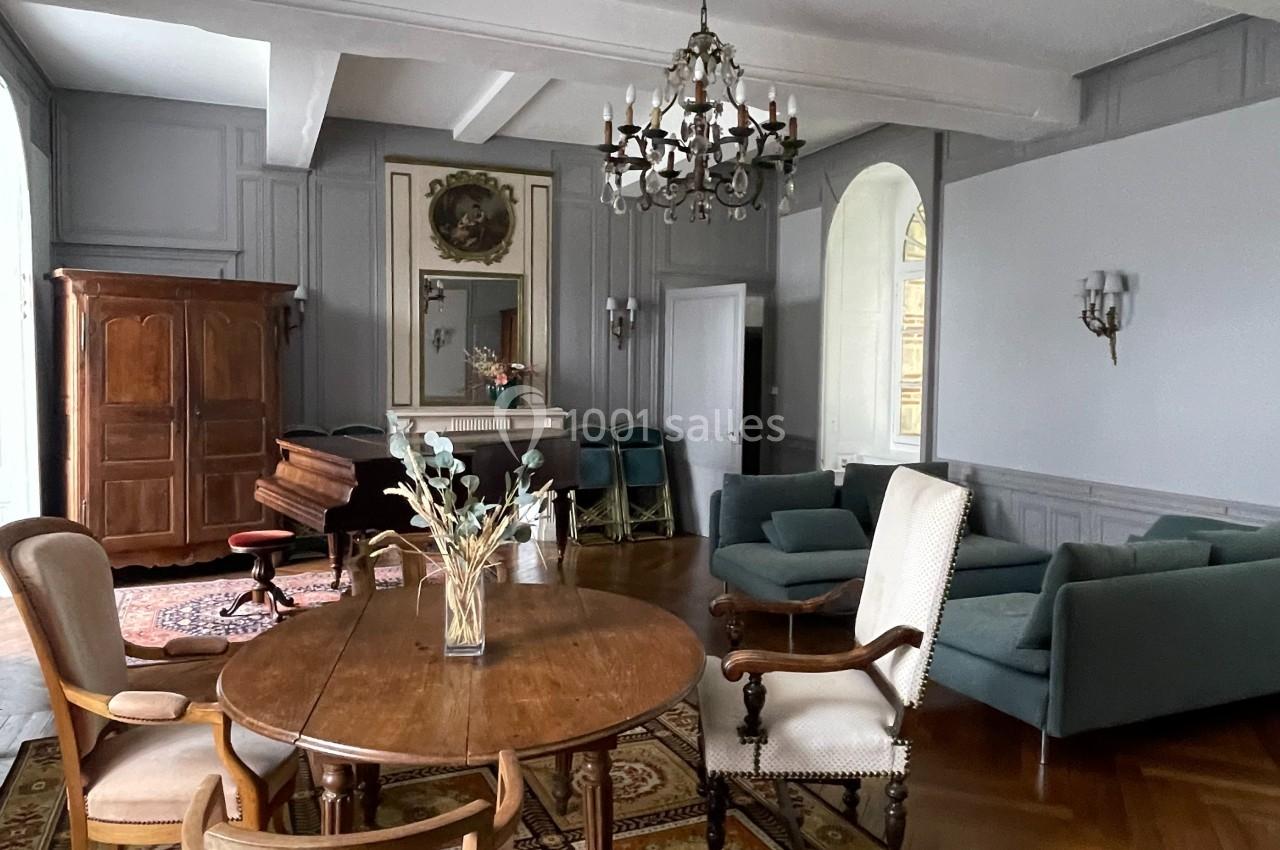Salle de séjour lumineuse avec mobilier ancien, tapis coloré, table en bois, fauteuils et lustre suspendu.