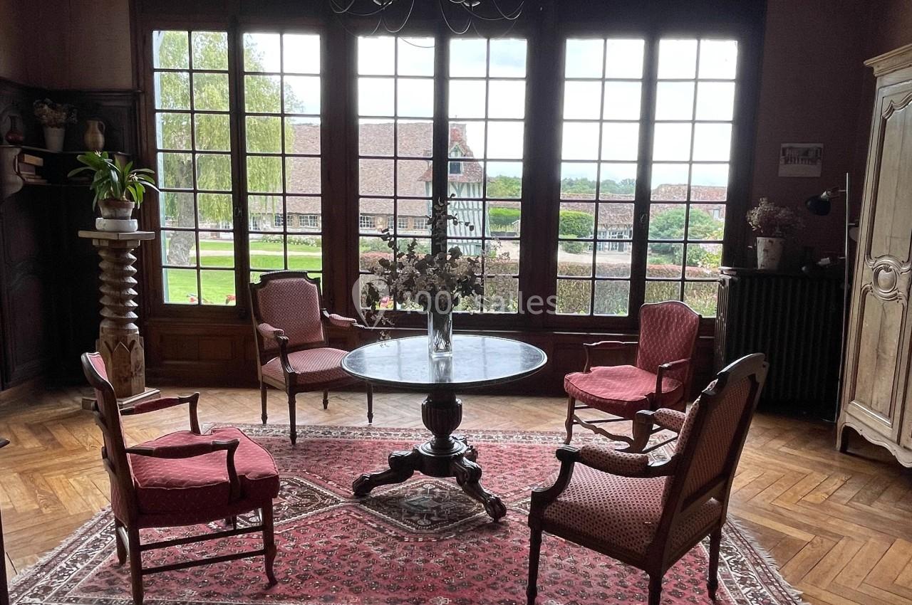 Salon avec parquet en bois, table ronde, quatre fauteuils rouges, tapis, chandelier et grandes fenêtres donnant sur un…