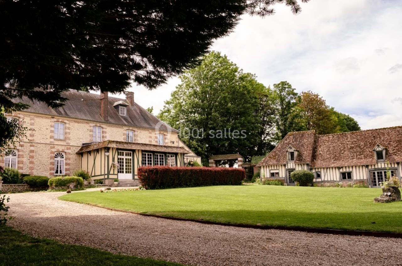 Manoir en pierre et maison à colombages entourés de pelouses, arbres et allée gravillonnée sous un ciel partiellement…