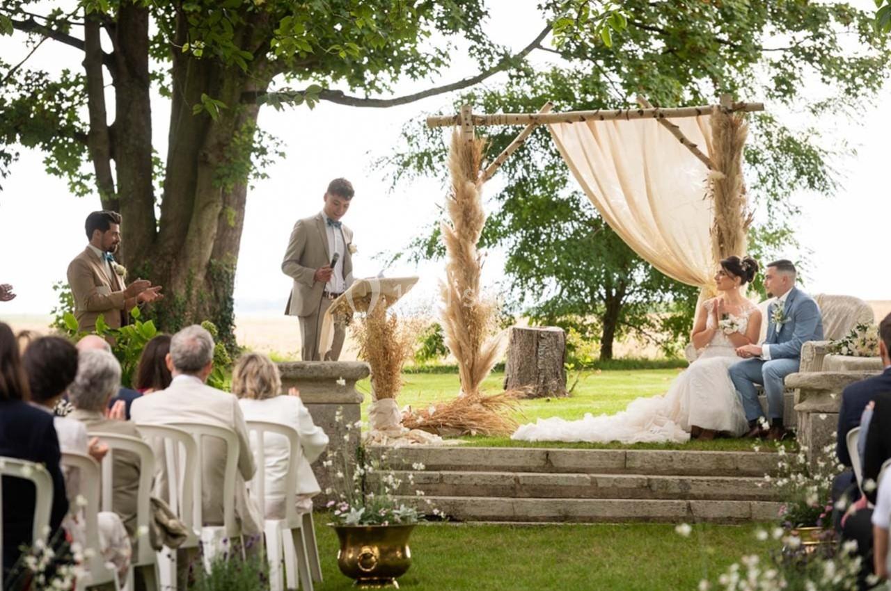 Un homme prononce un discours devant un couple assis sous une arche décorée, lors d'une cérémonie en plein air.