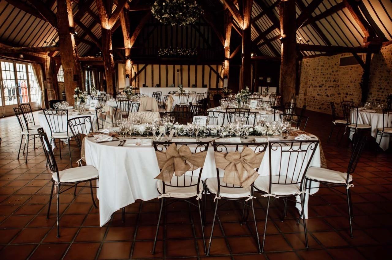 Salle de réception avec tables décorées, chaises ornées de nœuds en toile de jute et poutres apparentes en bois.