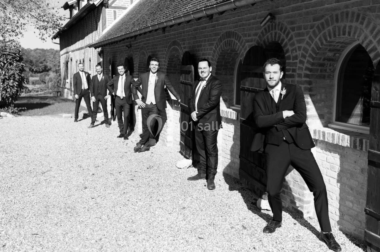 Six hommes en costume posent en ligne devant un bâtiment en briques avec des arches, sur une allée gravillonnée.