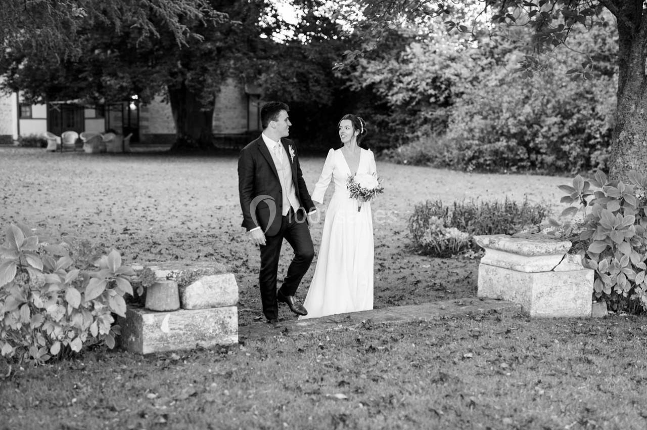Un couple en tenue de mariage marche dans un jardin, entouré de verdure et de pierres anciennes.