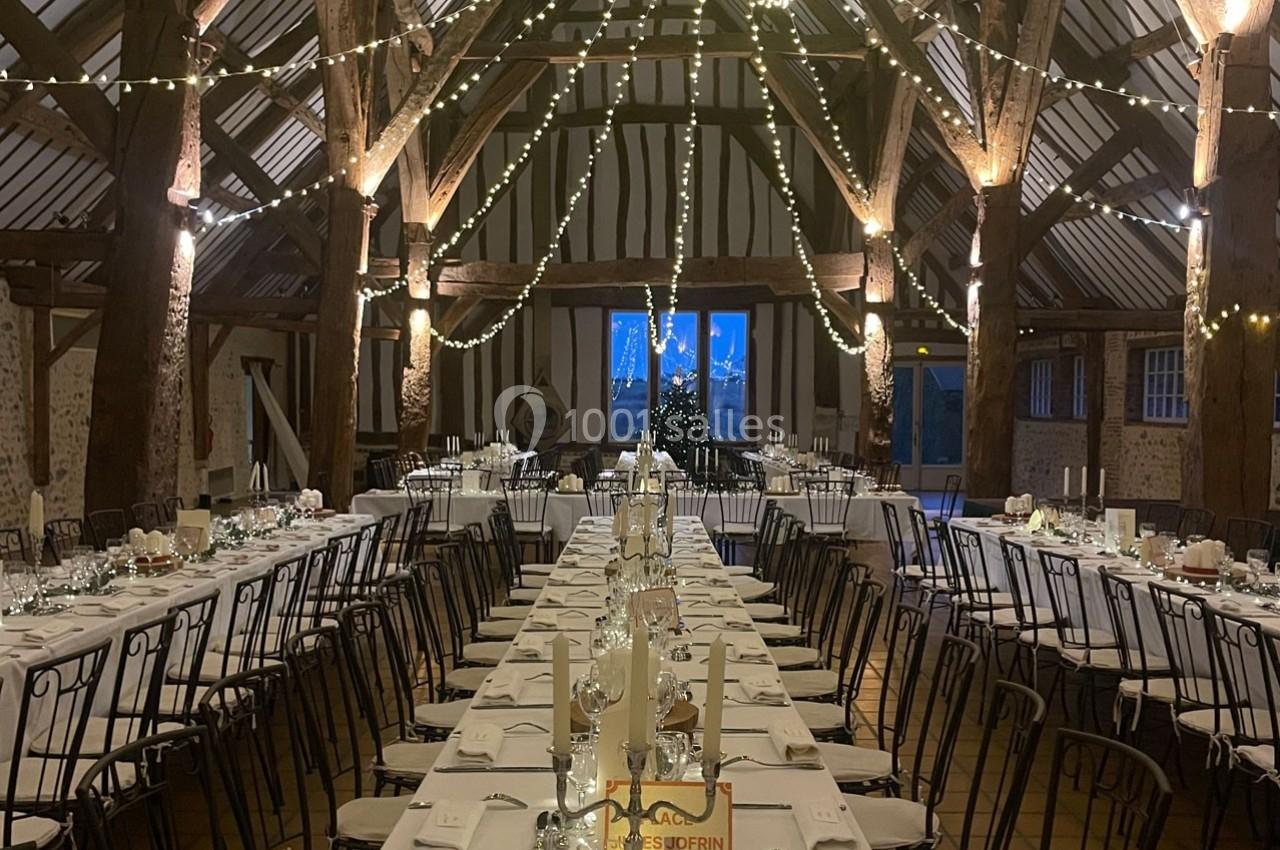 Grande salle de réception rustique avec poutres apparentes, tables dressées et guirlandes lumineuses suspendues.
