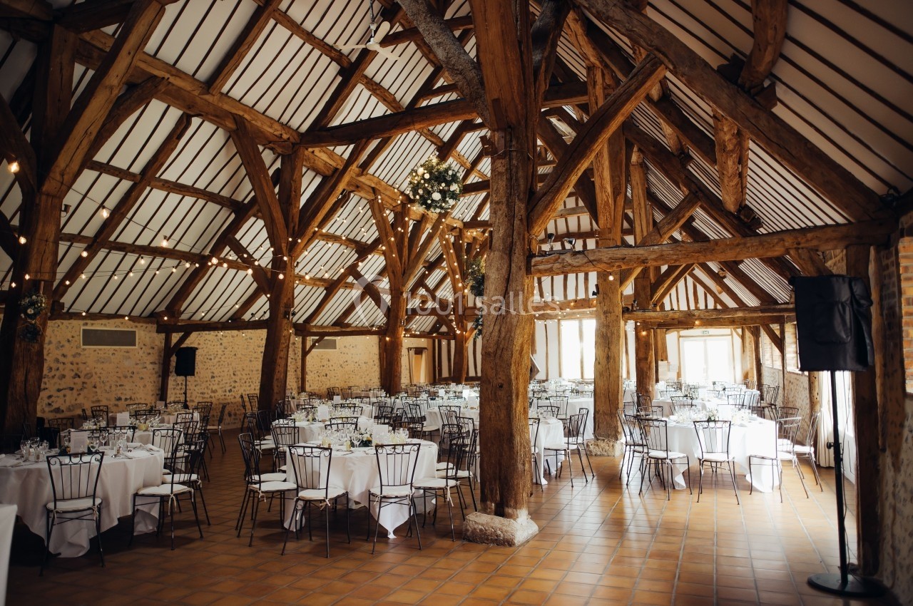 Salle de réception rustique avec poutres en bois apparentes, tables rondes dressées et lumière naturelle.