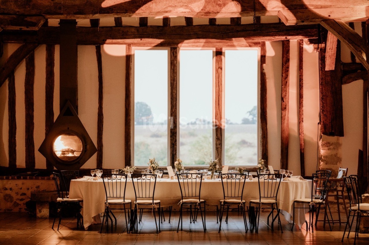 Grande table dressée avec des chaises en métal dans une salle rustique, éclairée par une cheminée et une lumière chaleureuse.