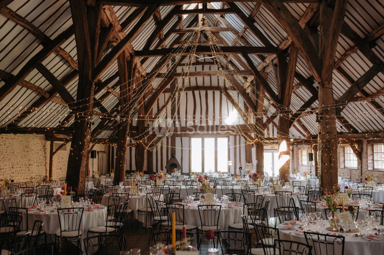 Salle de réception rustique avec poutres apparentes, tables rondes décorées et guirlandes lumineuses suspendues.