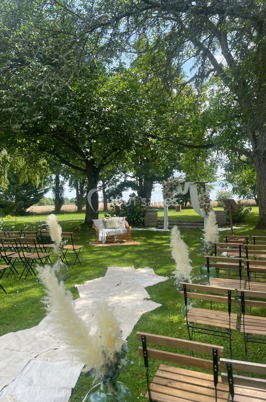 Allée en tissu bordée de bancs en bois et de décorations végétales, menant à une arche de mariage dans un jardin verdoyant.