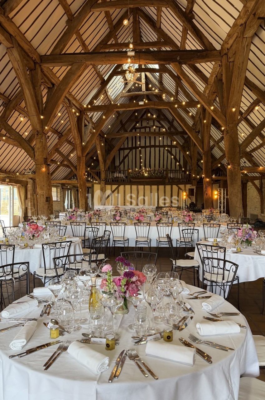 Grande salle de réception avec charpente en bois, tables rondes dressées avec nappes blanches et décorations florales.