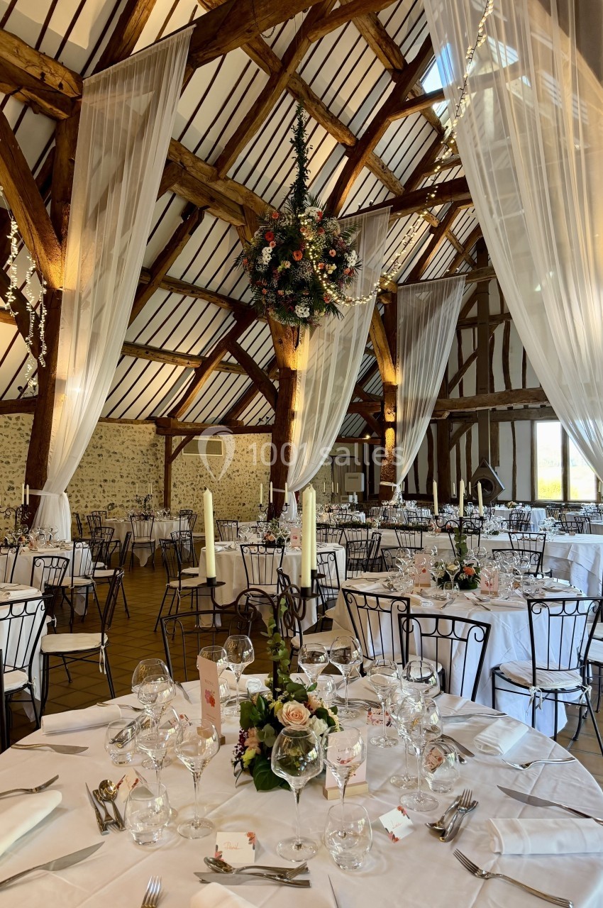 Salle de réception rustique avec poutres apparentes, tables rondes dressées et décoration florale élégante.