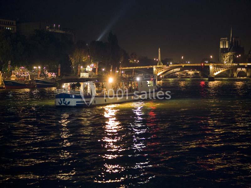Bateau illuminé naviguant sur la Seine la nuit, avec un pont et des bâtiments éclairés en arrière-plan.