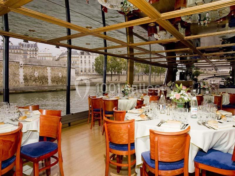 Salle de restaurant élégante avec vue sur une rivière, tables dressées avec nappes blanches et chaises en bois.