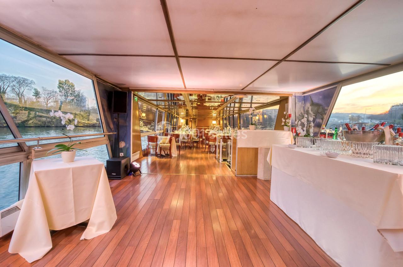 Intérieur d'un bateau avec parquet en bois, tables dressées et vue sur la rivière à travers de grandes baies vitrées.