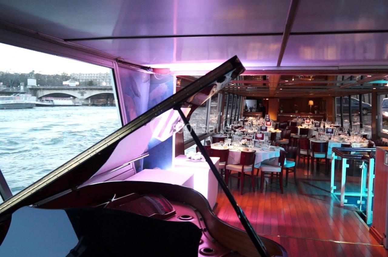 Intérieur d'un bateau avec un piano à queue au premier plan et une salle de dîner élégamment dressée en arrière-plan.