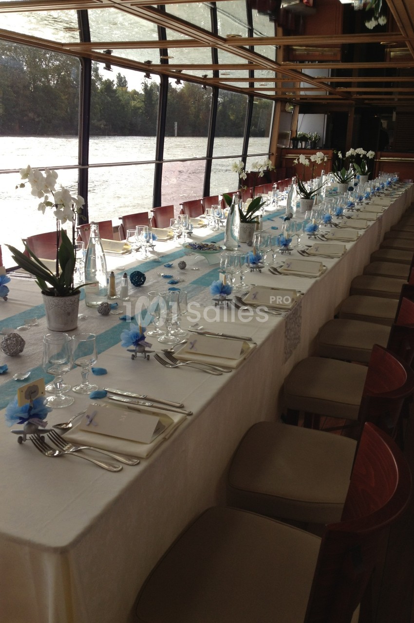Table élégamment dressée avec nappes blanches, décorations bleues et blanches, et orchidées, dans une salle avec vue sur l…