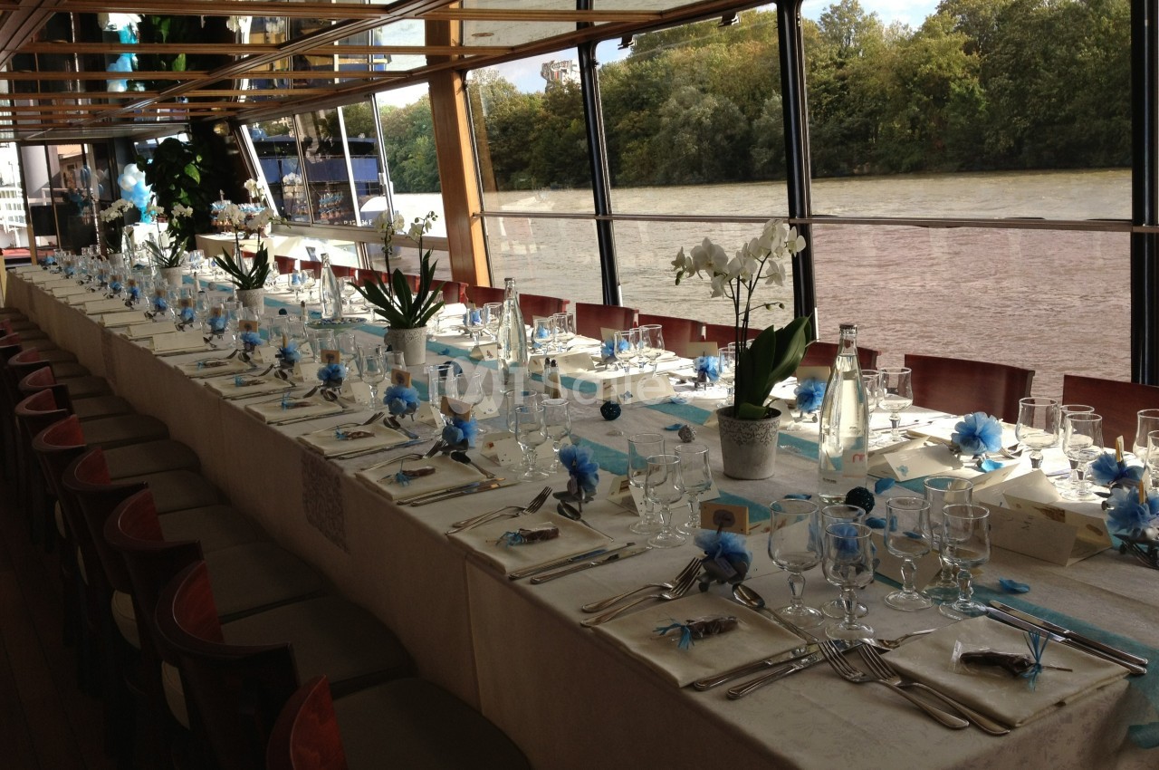 Table élégamment dressée avec nappes blanches, décorations bleues et vue sur une rivière à travers de grandes baies vitrées.