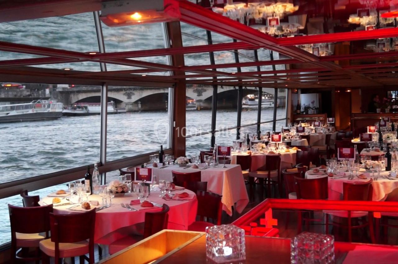 Salle de restaurant avec tables dressées, grandes baies vitrées donnant sur une rivière et pont en arrière-plan.