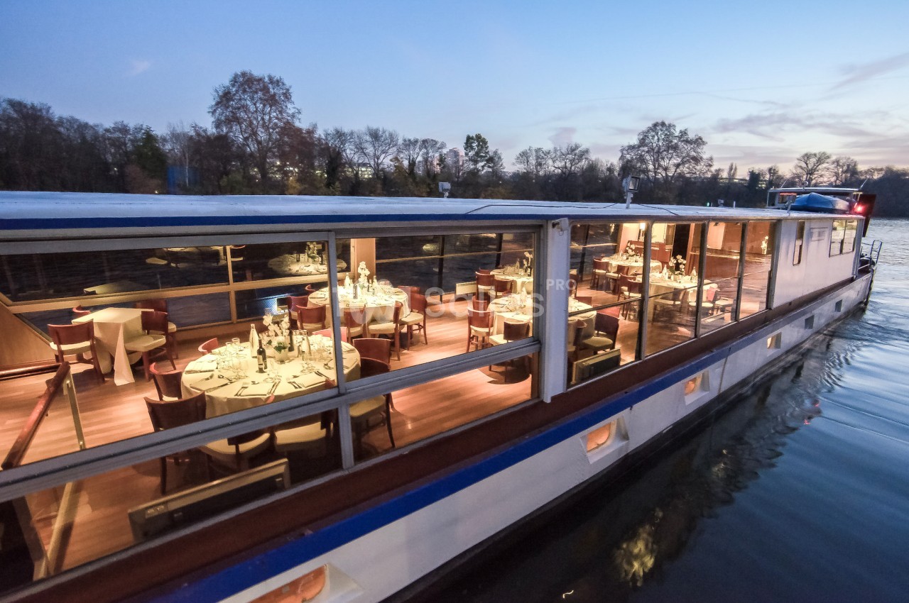 Bateau-restaurant éclairé naviguant sur un fleuve au crépuscule, avec des tables dressées visibles à travers les baies…