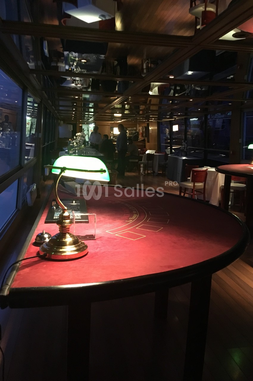 Table de jeu rouge éclairée par une lampe verte dans une salle sombre avec des fenêtres et des tables en arrière-plan.
