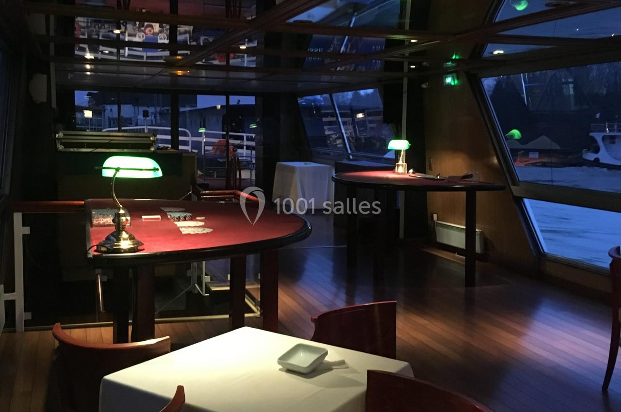 Salle intérieure d'un bateau avec des tables en bois, nappes blanches, éclairage tamisé et vue sur l'extérieur.