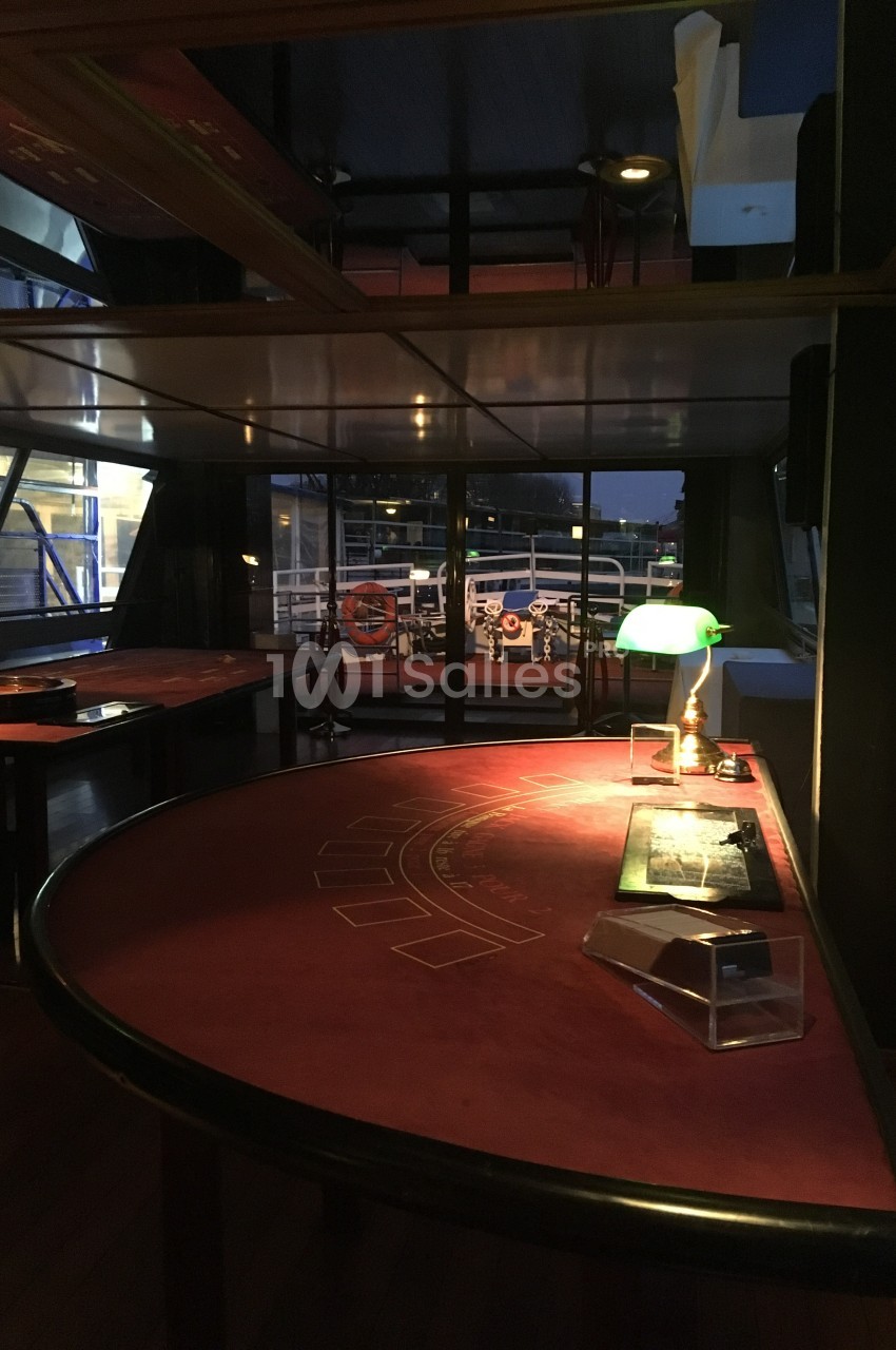Salle avec une table de jeu rouge, une lampe verte allumée et une vue sur l'extérieur à travers des baies vitrées.