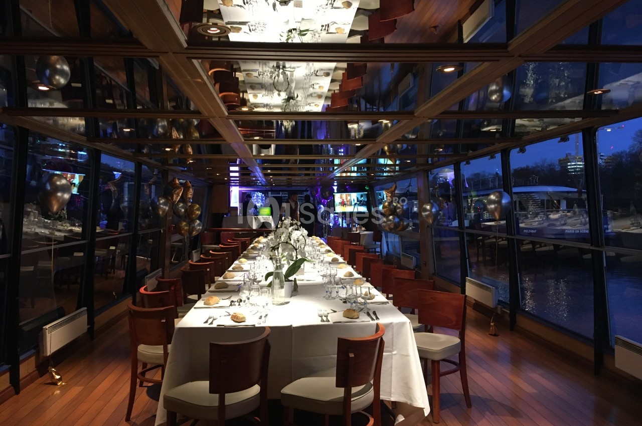 Salle de réception élégante avec une longue table dressée, entourée de chaises en bois, et vue sur l'extérieur vitré.