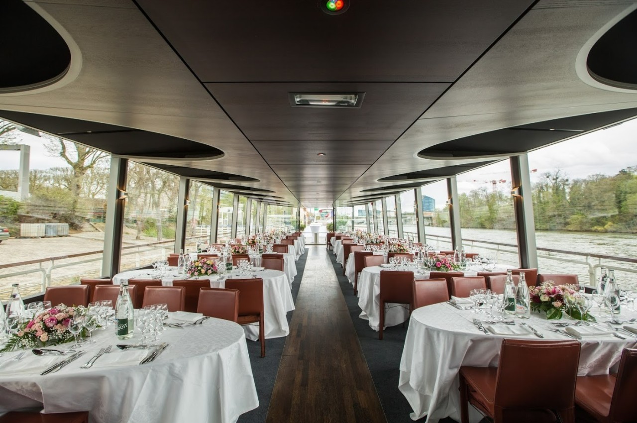 Salle de réception sur un bateau avec des tables rondes dressées, entourées de chaises en cuir, et vue sur un paysage…