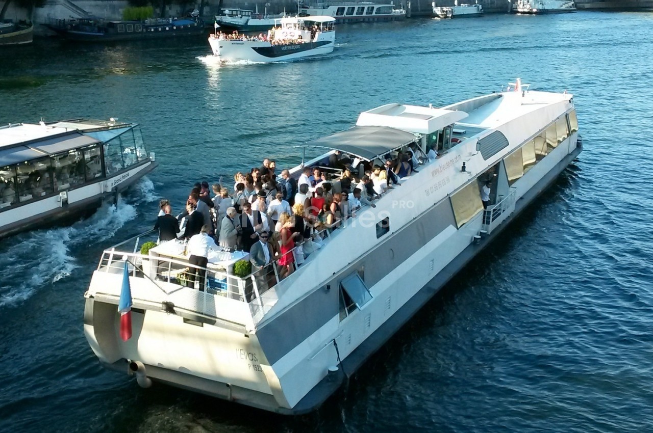 Bateau de croisière rempli de passagers naviguant sur une rivière, avec d'autres embarcations en arrière-plan.