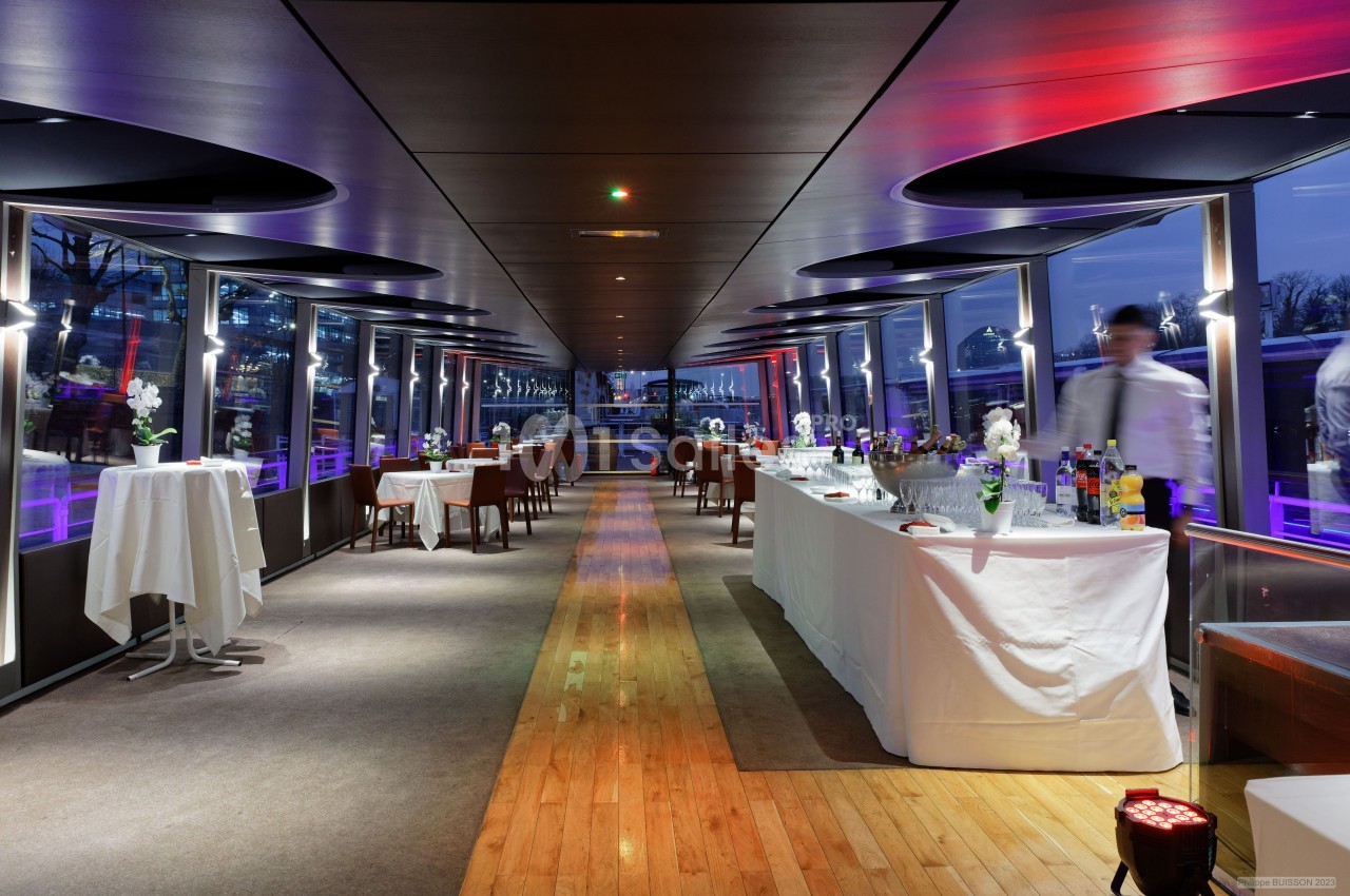 Salle moderne avec baies vitrées, tables dressées, buffet et serveur en mouvement, éclairage tamisé.