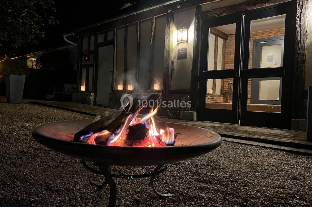 Brasero allumé dans une cour gravillonnée devant un bâtiment éclairé avec des murs en bois et des portes vitrées.
