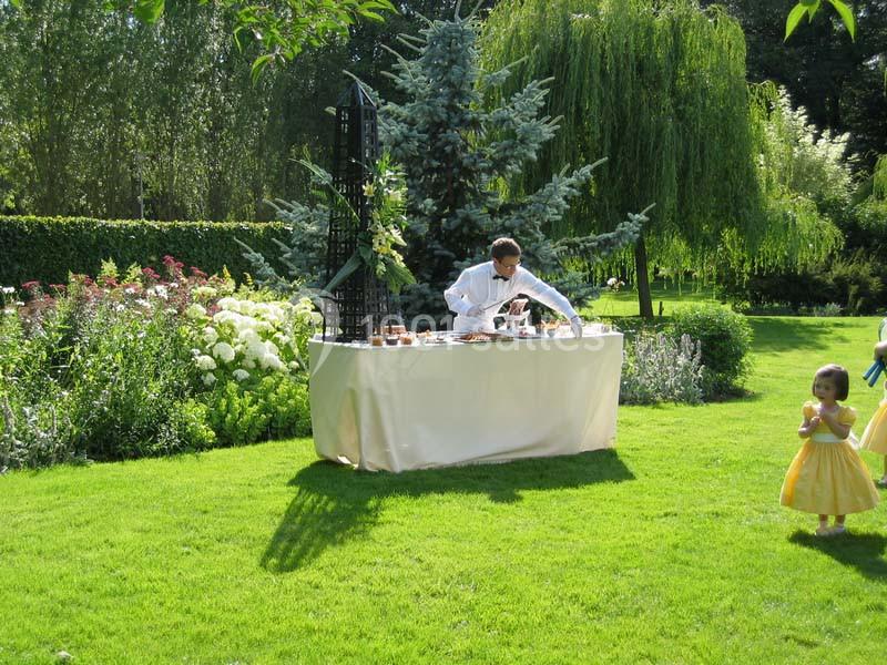 Un homme prépare une table dressée dans un jardin verdoyant, avec des fleurs et des arbres en arrière-plan.