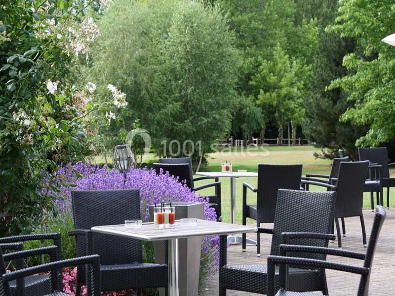 Terrasse avec mobilier en rotin noir, table dressée avec verres, entourée de fleurs et d'arbres dans un jardin verdoyant.