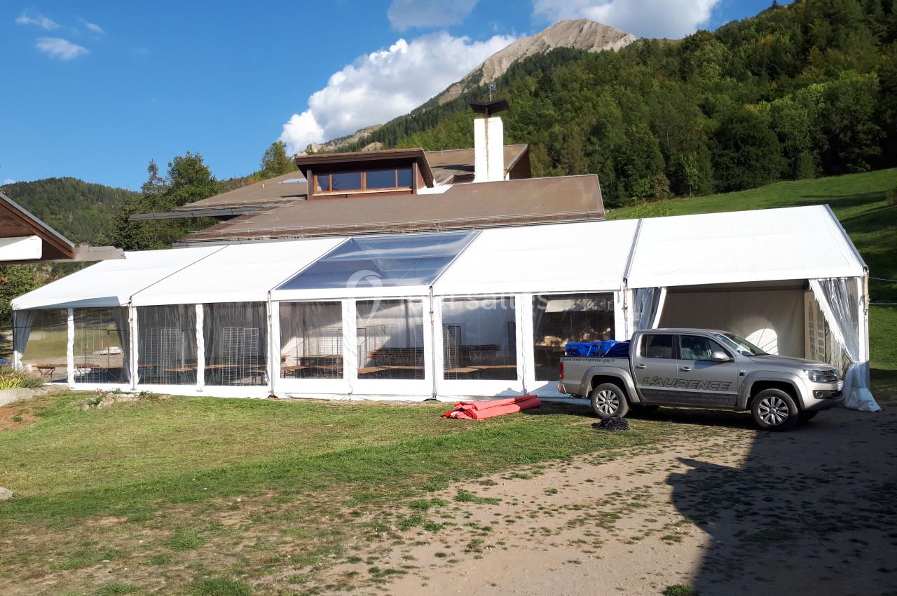 Grande tente blanche installée sur une pelouse près d'une maison en bois, avec un pick-up stationné à côté.