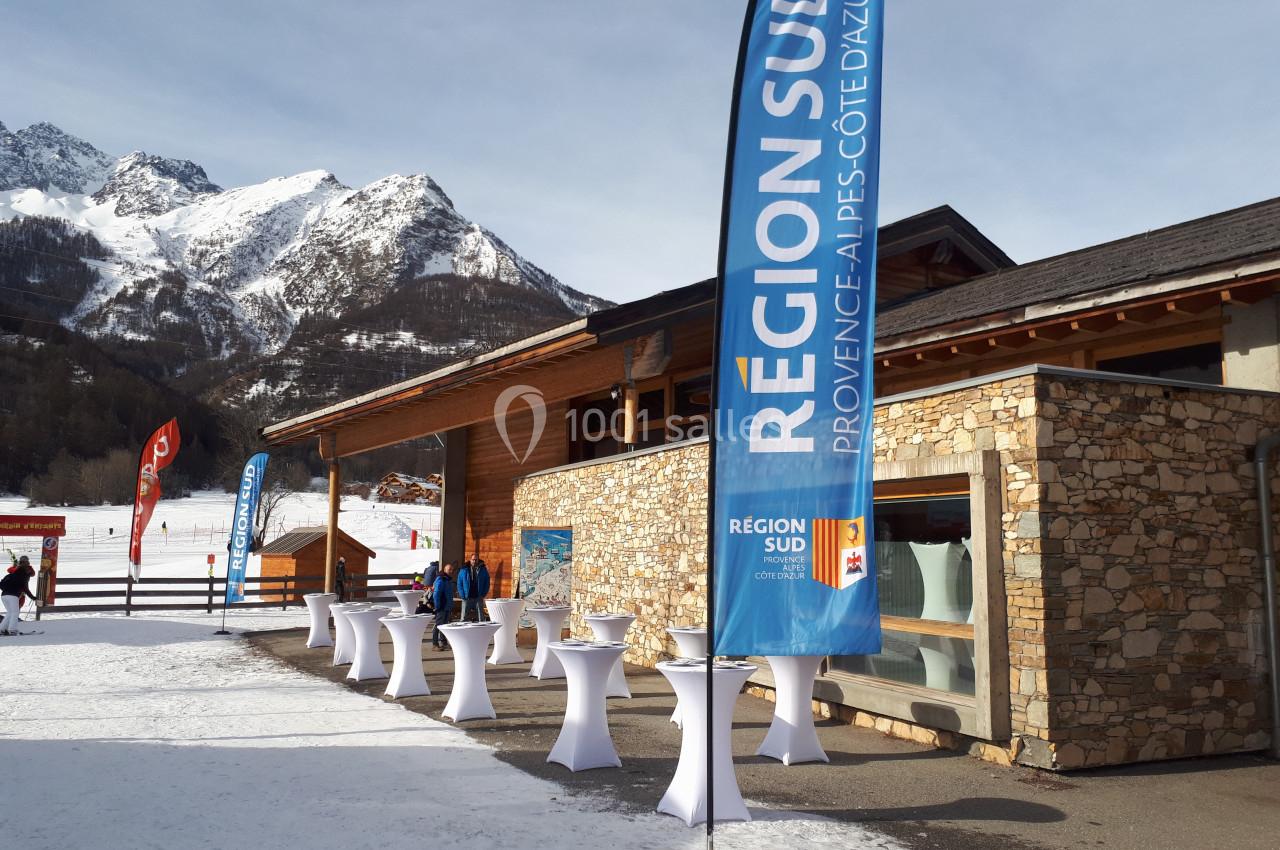 Bâtiment en pierre et bois devant des montagnes enneigées, avec des drapeaux ’Région Sud’ et des tables hautes installées.