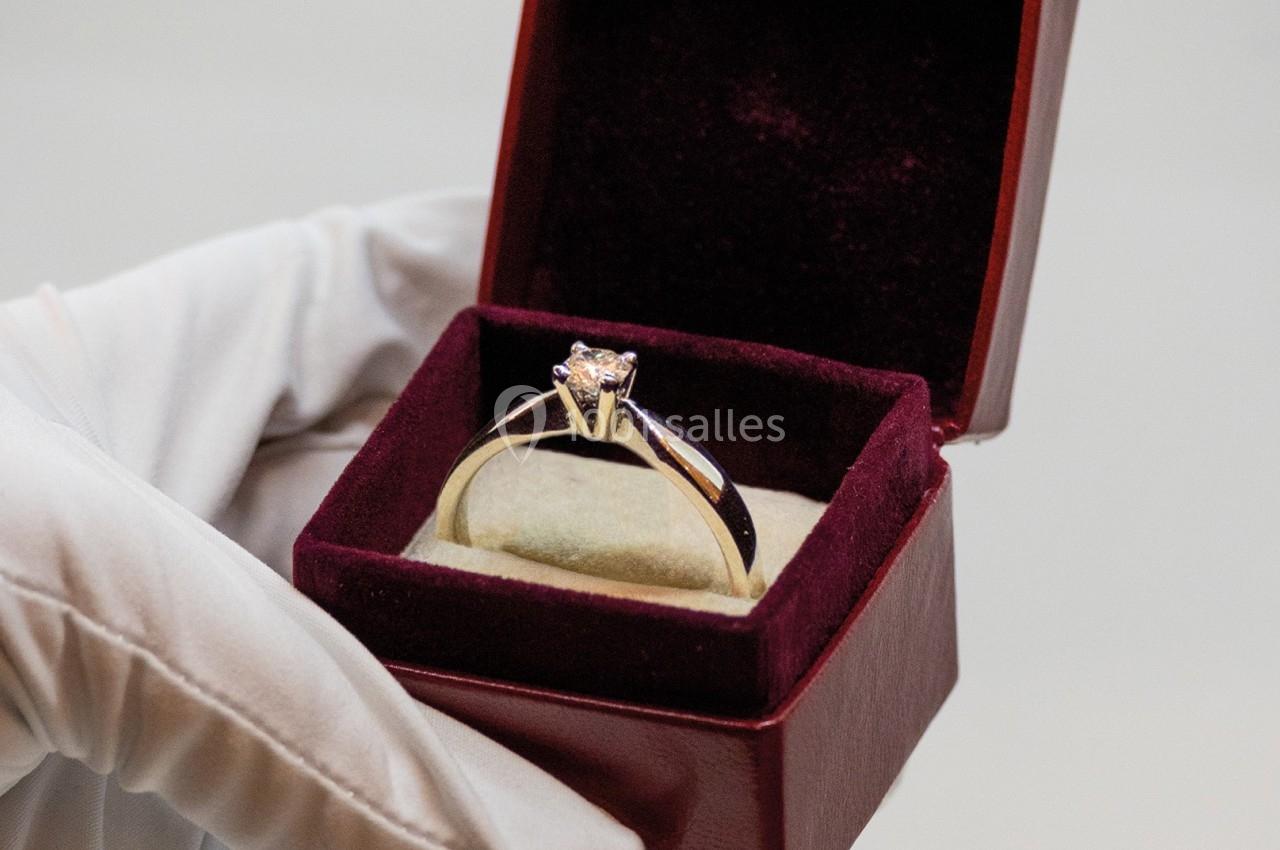 Bague en or avec un diamant, présentée dans un écrin rouge ouvert, tenue par une main gantée.