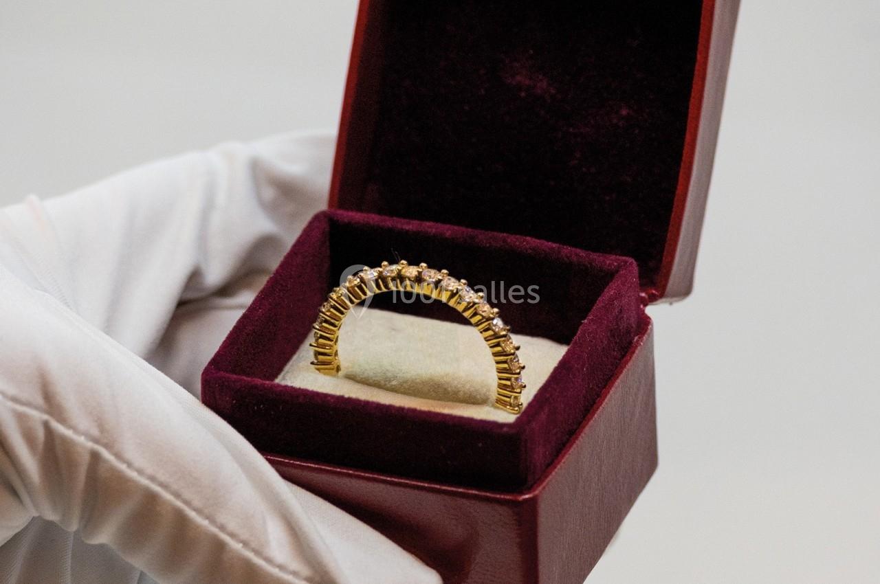 Une main gantée tient un écrin ouvert contenant une bague en or ornée de petites pierres.