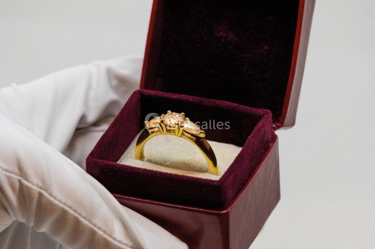 Bague en or ornée de pierres précieuses dans un écrin rouge, tenue par une main gantée blanche.