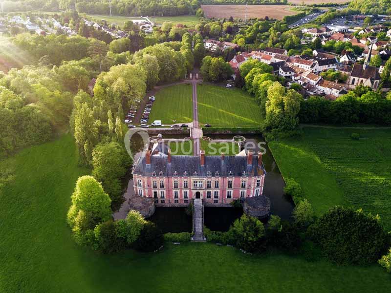 Vue aérienne d'un château entouré de douves, avec un parc verdoyant et un village en arrière-plan.