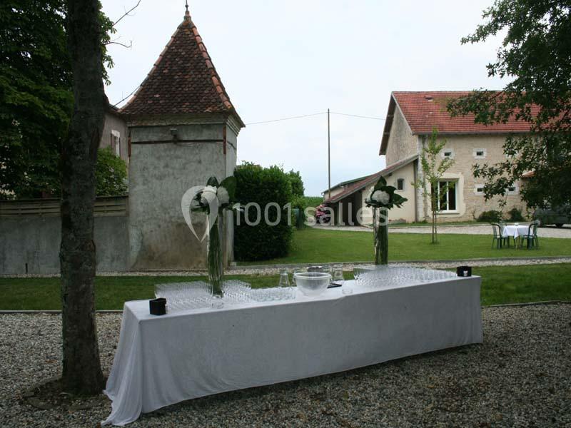 Table dressée en extérieur avec nappage blanc, verres et décorations florales, dans une cour avec bâtiments en arrière-plan.